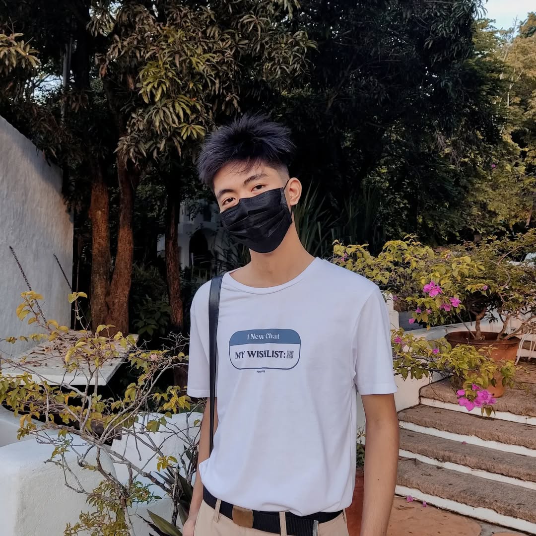 Jhestin in a white T-shirt and black face mask, standing among trees and bougainvillea.